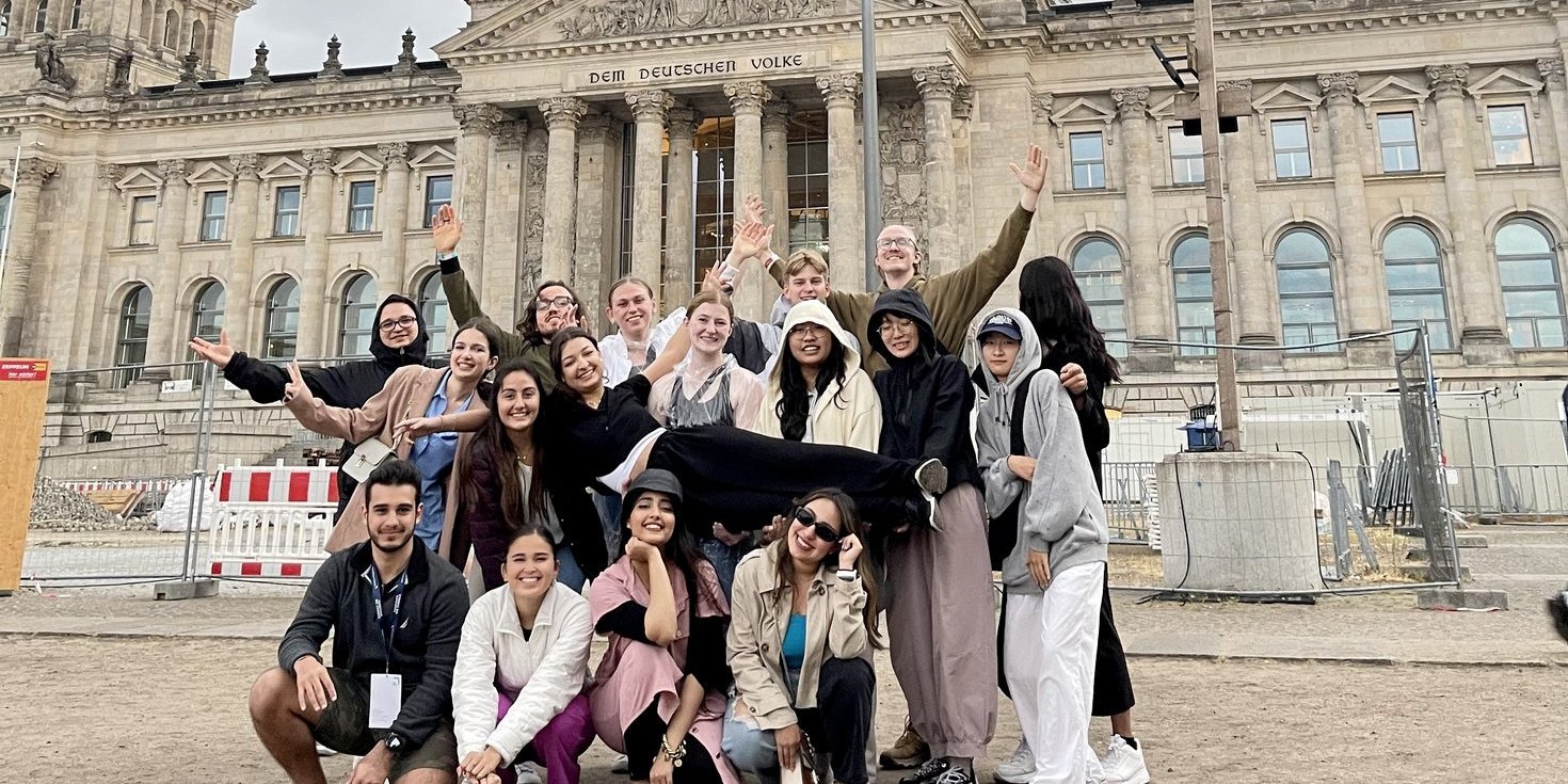 A group of international students is posing in fron of the German parliament building in Berlin. The German flag can be seen in the background. 