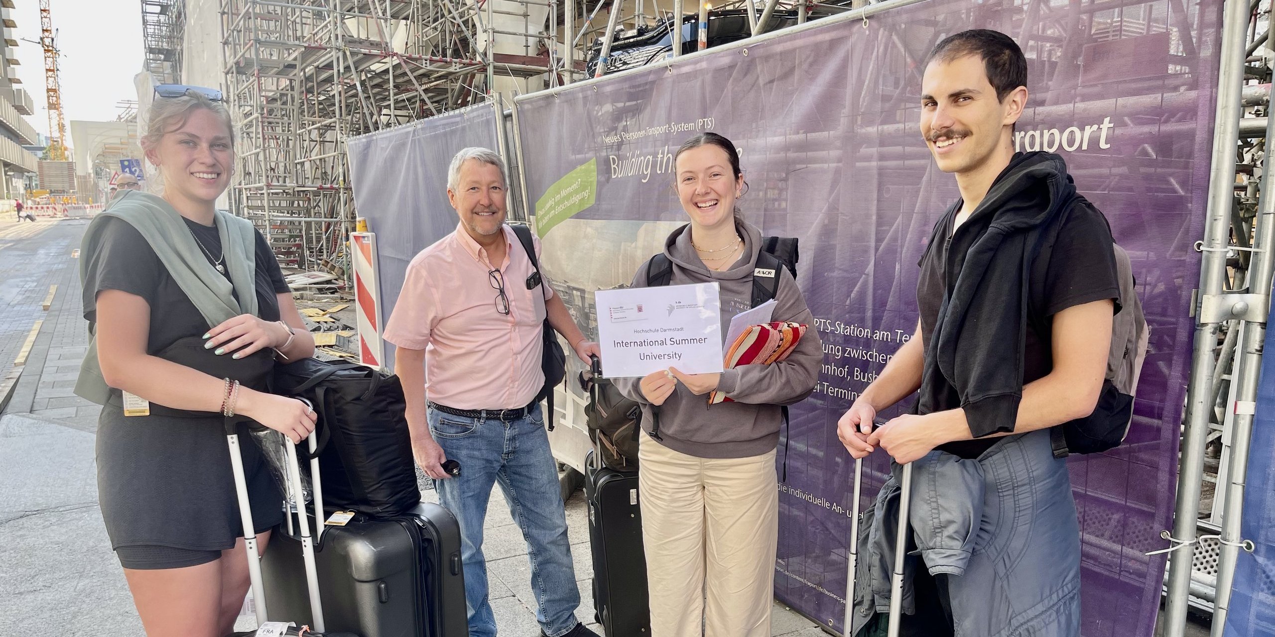 Two international students and a professor are holding their suitcases and are beeing greeted by a student assistant at the Frankfurt Main International Airport.  Zwei internationale Studierende und ein Professor stehen mit ihren Koffern zusammen und werden am Frankfurter Flughafen von einer studentischen Hilfskraft willkommen geheißen. 